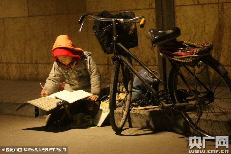 鄭州中原路京廣鐵路大橋下涵洞的馬路牙子邊，一位戴著紅色辣椒帽的小男孩趴在一個(gè)小馬扎上，上面鋪著一張車墊，在微弱的燈光下寫作業(yè)