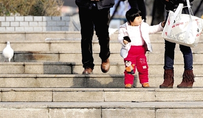 昨天中午，一女孩拉著家長的手在紫荊山公園曬太陽，氣溫升高，孩子的棉衣解開了扣子。